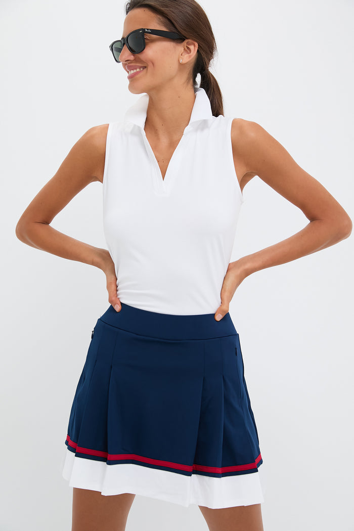 A woman stands confidently with hands on hips, wearing Tnuck Sport sunglasses, a white sleeveless Caroline polo, and a navy blue pleated skirt featuring red and white stripes.