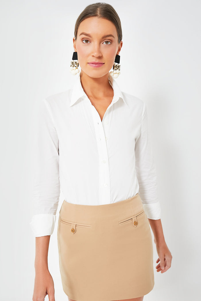 A woman wears a white cotton poplin STAUD bodysuit with rolled sleeves and a beige skirt, standing confidently.