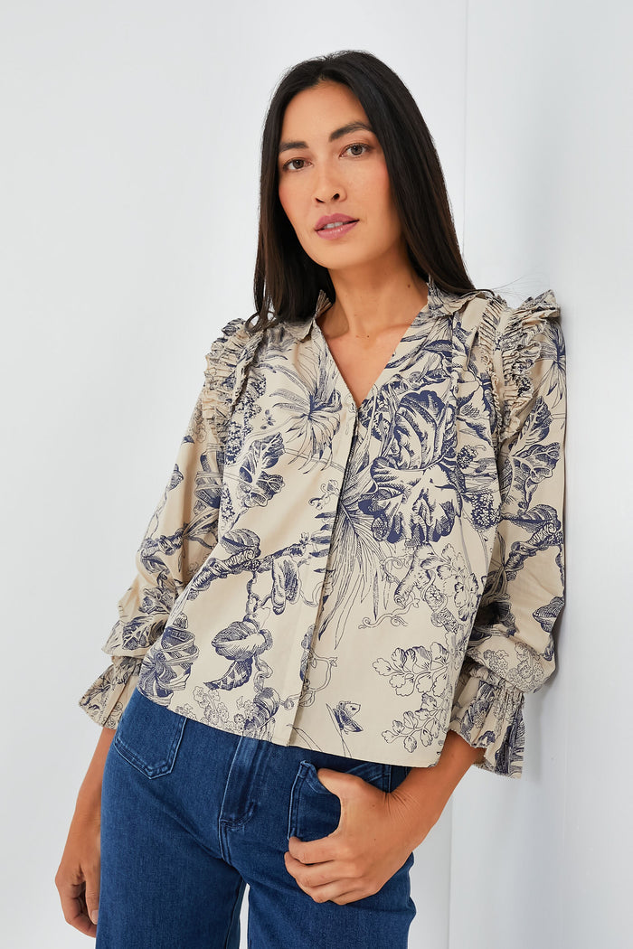 A woman with long dark hair wears a Love the Label Jessie Top, a beige blouse with dark floral print and ruffled details, paired with blue jeans.