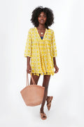 A woman in a Ro's Garden yellow patterned vacation-ready V-neck tunic with tassels holds a woven tote and wears flat sandals.