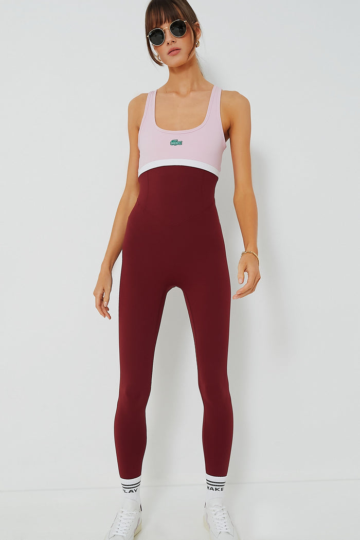 A woman in sunglasses wears a pink and burgundy sleeveless colorblocked one-piece jumpsuit by Bandier x Lacoste, paired with white sneakers and socks, showcasing stylish tennis performance wear.